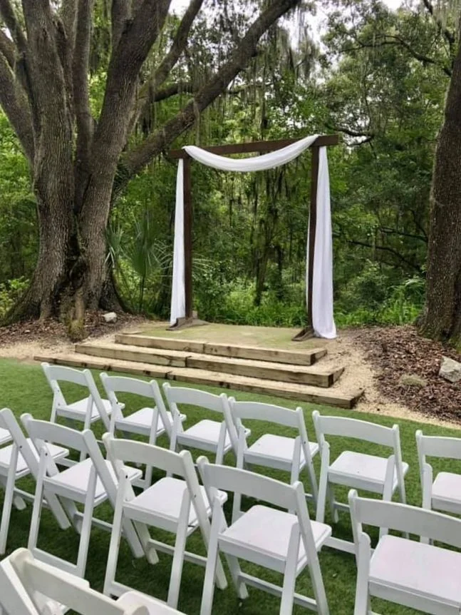 Outdoor wedding ceremony setup with white chairs and an arch draped in white fabric among trees.