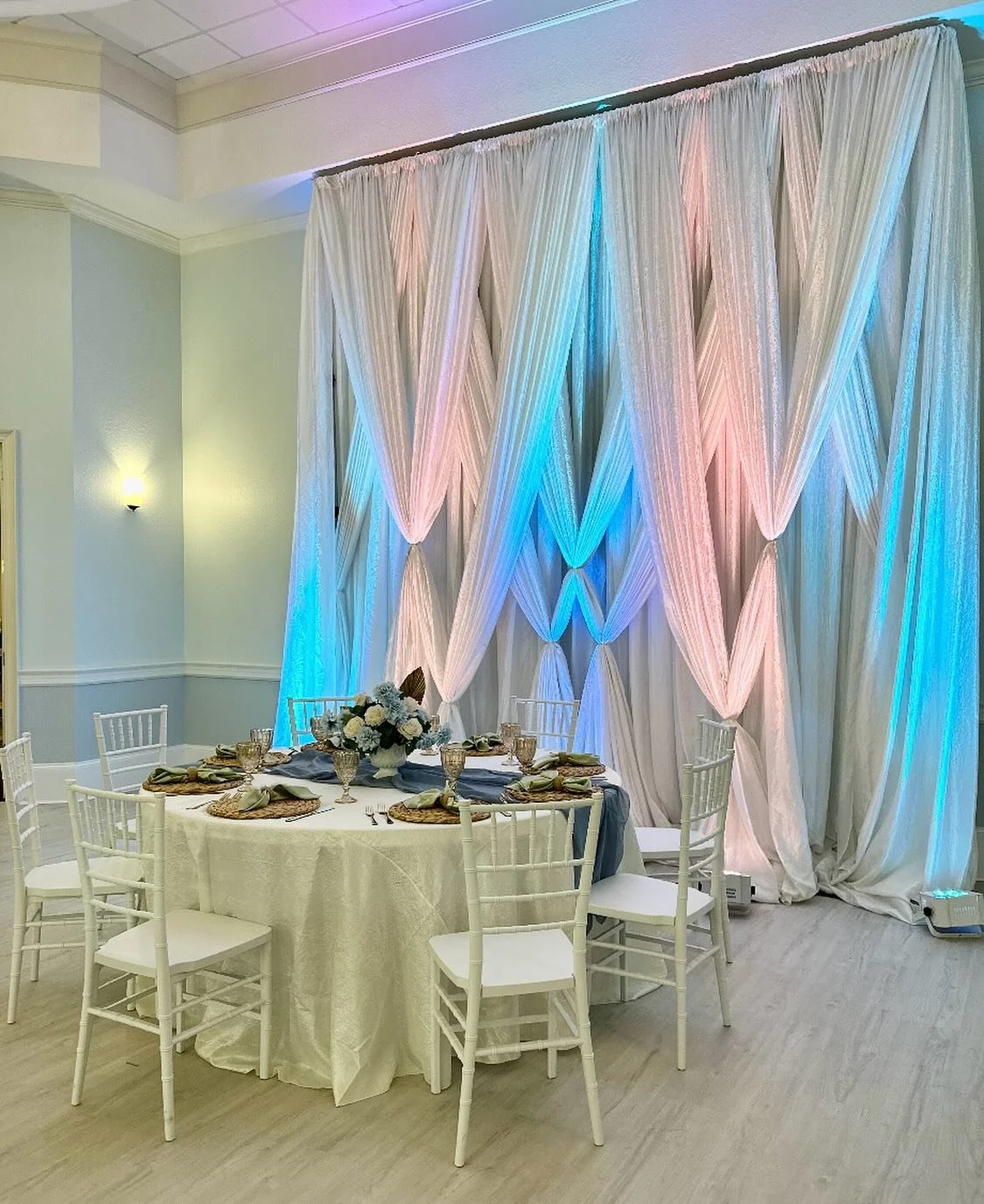 Draped Designed Backdrop, Up Lighting, White Chiavari Chairs, Vintage Styled Goblets, & Water Hyacinth Placemats. ——————————————————&mdash