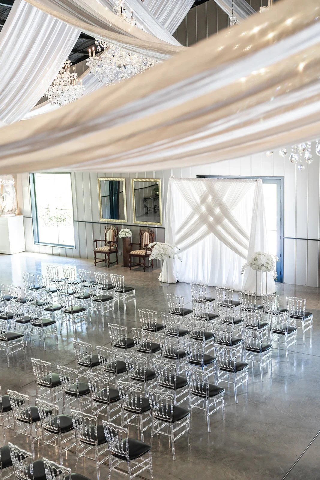 Wedding venue with clear chairs, white drapery, and chandeliers.
