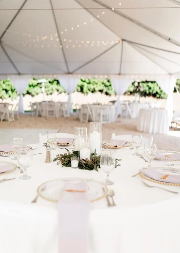 Outdoor wedding reception setup under a tent with white tablecloths, place settings, candles, and greenery centerpiece.