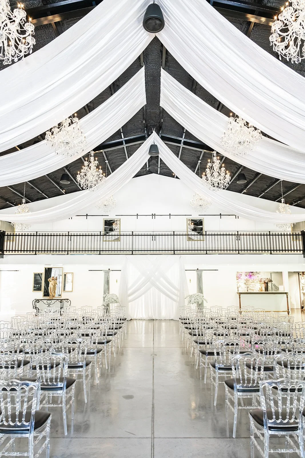 Elegant wedding venue with white drapery, chandeliers, and transparent chairs arranged for a ceremony.