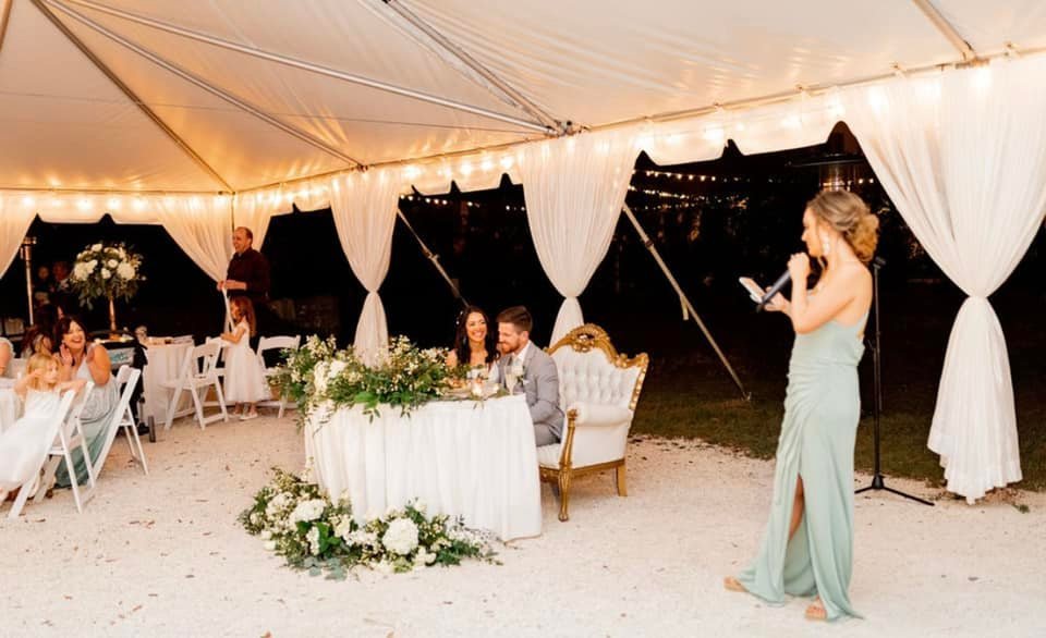 Wedding reception under a tent with people seated at tables, a couple sitting at a decorated sweetheart table, and a woman in a green dress speaking with a microphone.