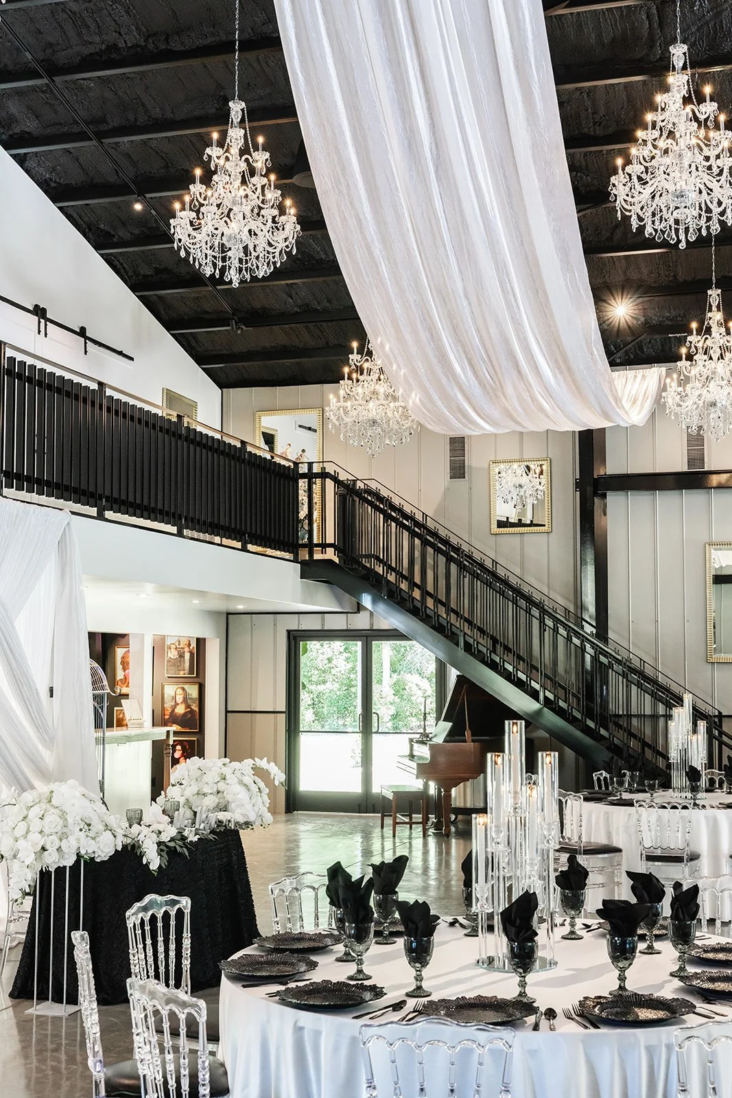 Elegant event space with chandeliers, draped white fabric, round tables set with black and white tableware, clear chairs, grand piano, and black staircase.