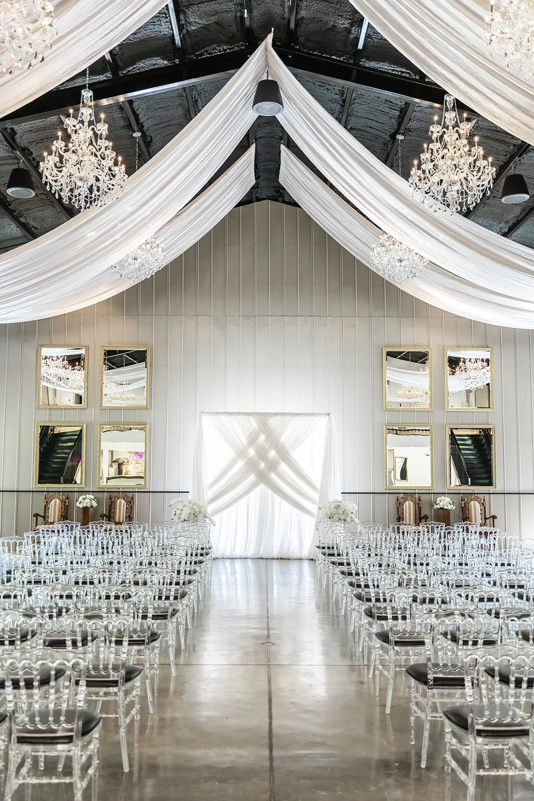 Elegant indoor wedding venue with white drapery ceiling, chandeliers, and rows of transparent chairs.