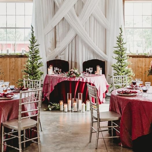 Elegant indoor wedding setup with round tables covered in red tablecloths, silver chairs, and a central sweetheart table adorned with candles and greenery. White drapes and small decorative trees enhance the rustic ambiance.