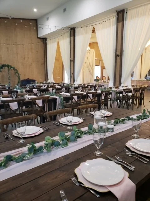 Elegantly set tables in a rustic venue with wooden chairs, white plates, pink napkins, and a greenery table runner. White drapes and soft lighting create an inviting atmosphere, suitable for a wedding or similar event.