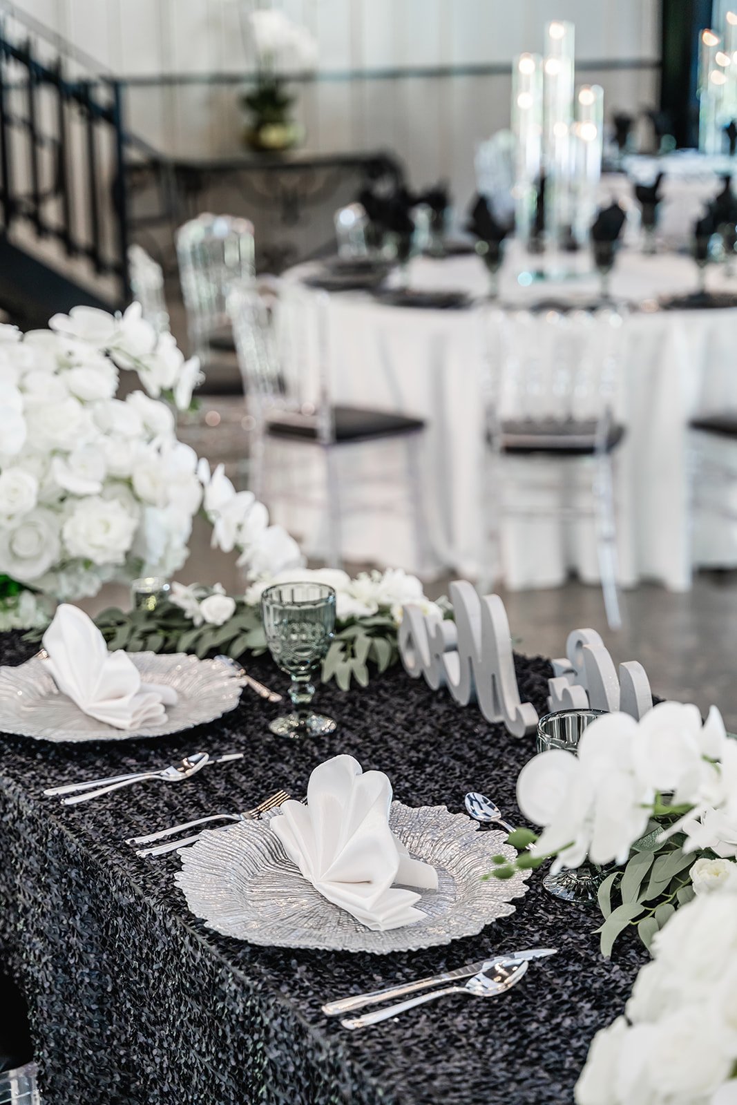 Elegantly set table with black tablecloth, glass plates, white napkins folded in a fan shape, greenery, white flower arrangements, and decorative elements, in a sophisticated dining setting.