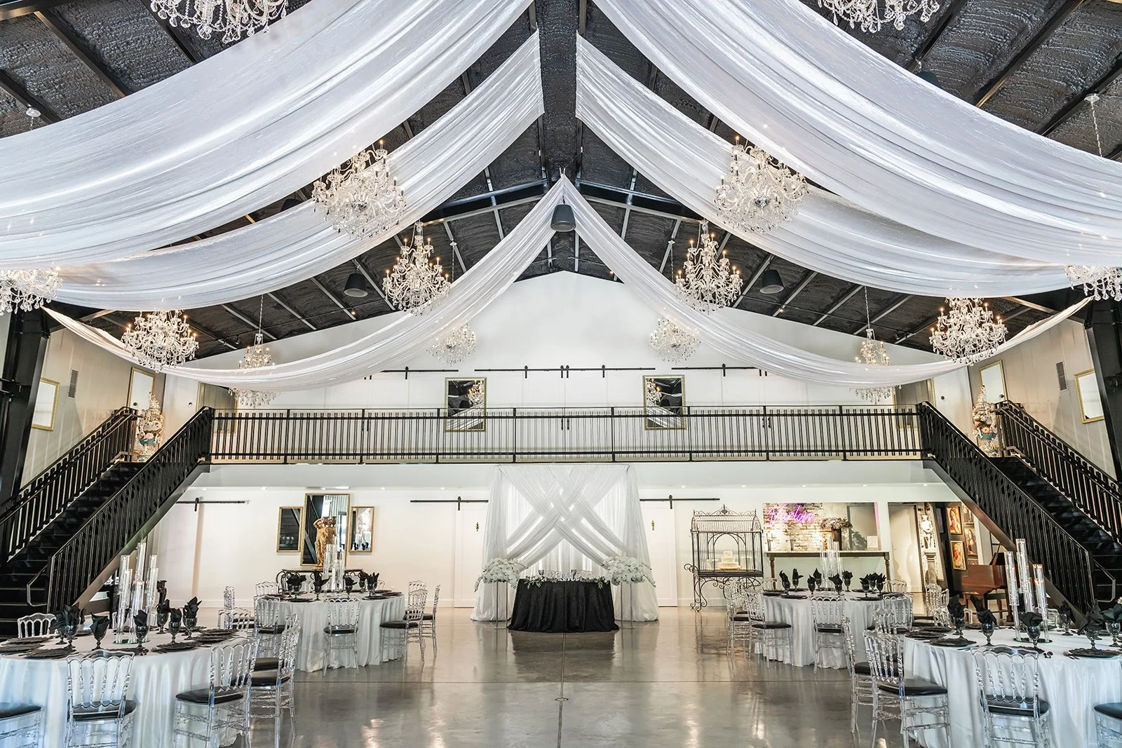 Elegant event venue interior with white draped ceiling, chandeliers, black staircase, and round dining tables with white tablecloths