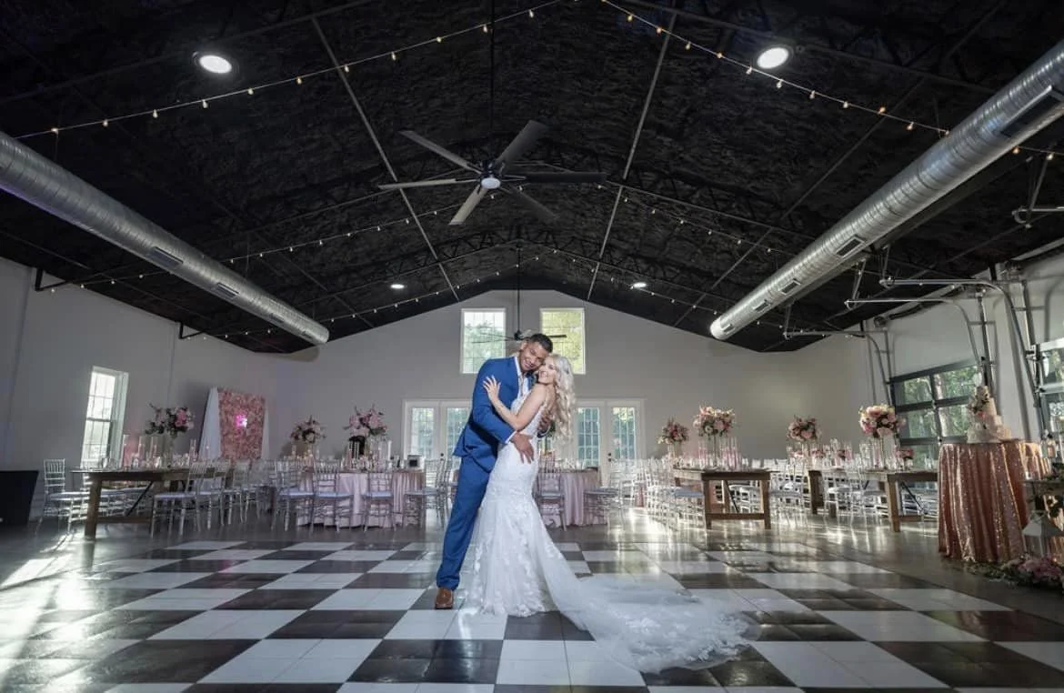 A couple in wedding attire embracing in a large venue with checkered flooring, overhead lighting, and decorated tables for a wedding reception.