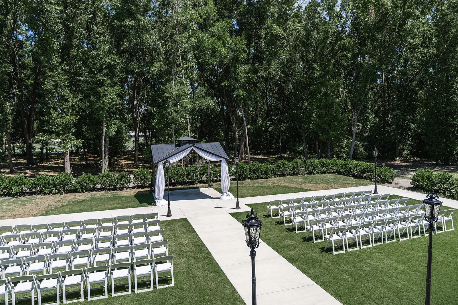 Outdoor wedding ceremony setup with white chairs and a decorative gazebo on a grassy area surrounded by trees.