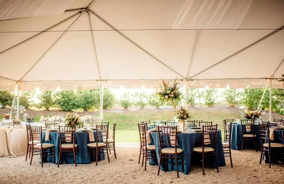 Outdoor wedding reception setup under a large white tent with round tables covered in blue tablecloths, adorned with floral centerpieces. Wooden chairs surround the tables, and there is soft lighting creating a warm ambiance.