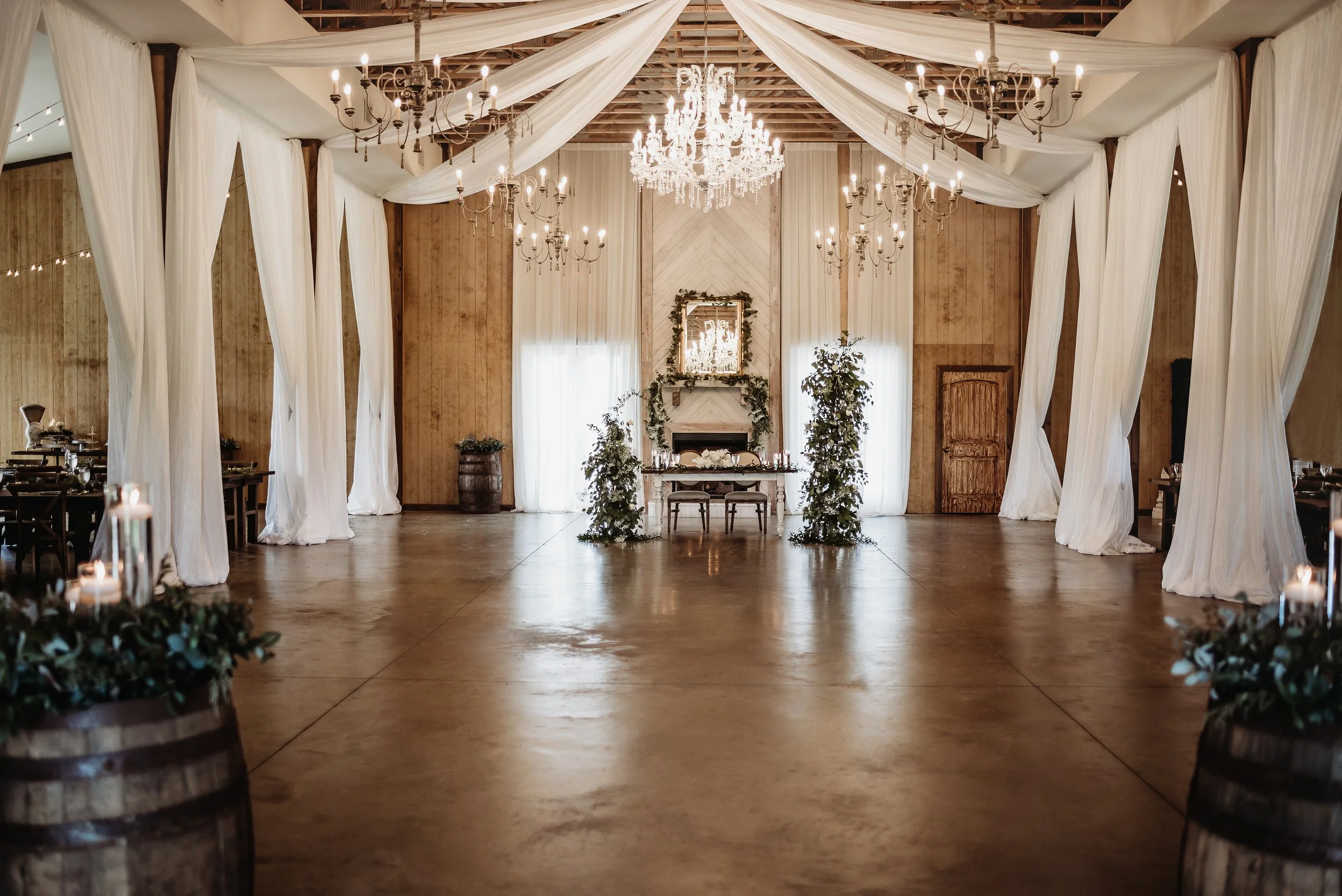 Elegant wedding venue with chandeliers, white drapery, and greenery. Centered table under a mirror with floral decorations. Wooden barrels with candles and foliage accent the space.