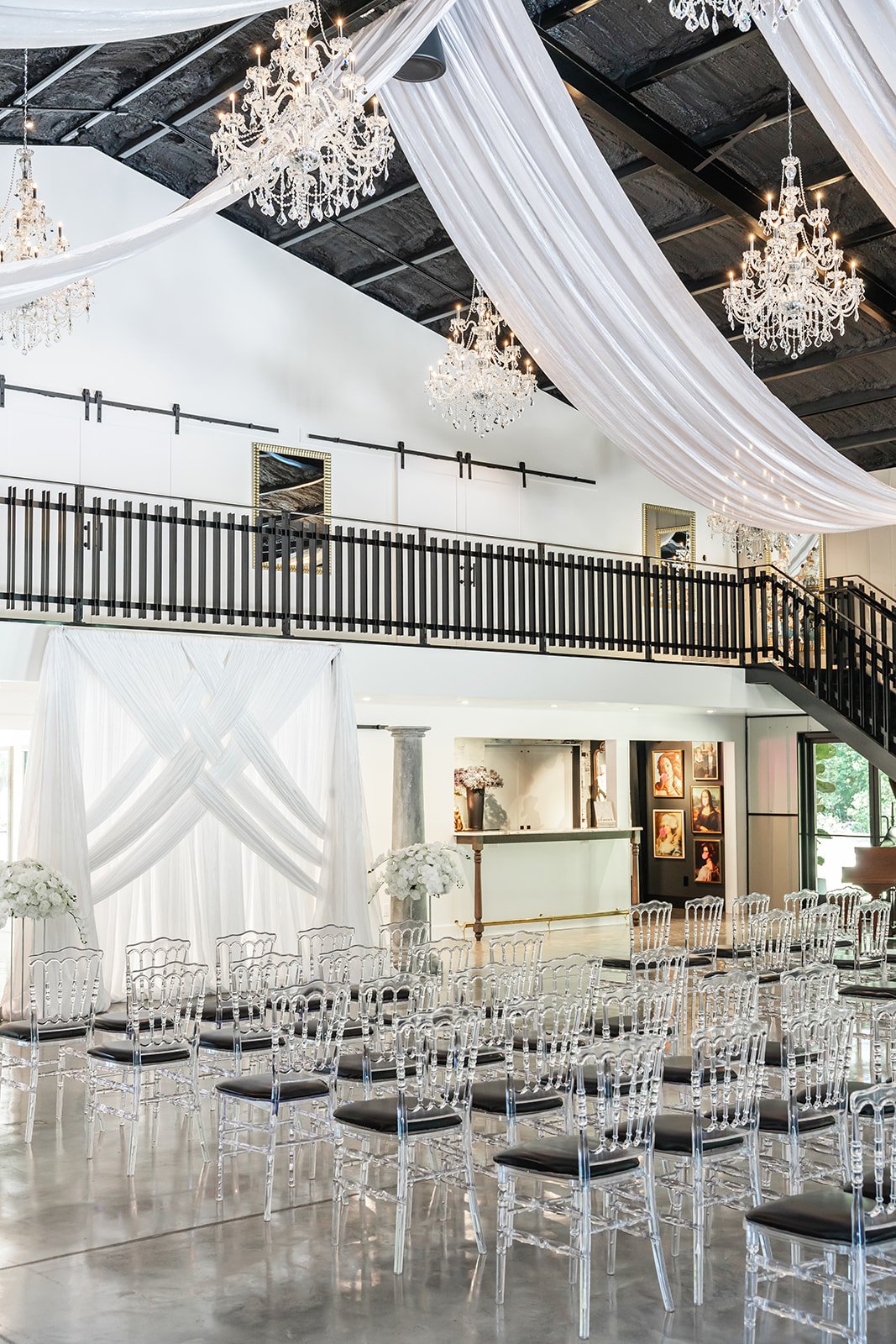 Indoor wedding venue with transparent chairs, white drapes, chandeliers, and a decorated altar area.