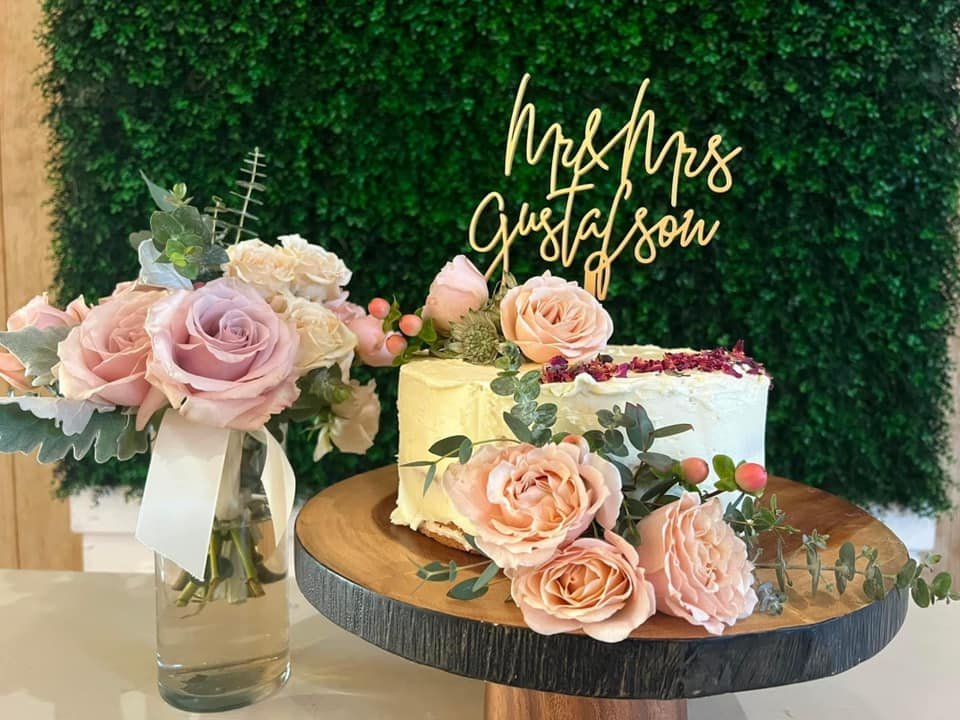 Wedding cake with "Mr & Mrs Gustafson" topper, surrounded by pink roses and greenery on a wooden stand, next to a bouquet in a glass vase.