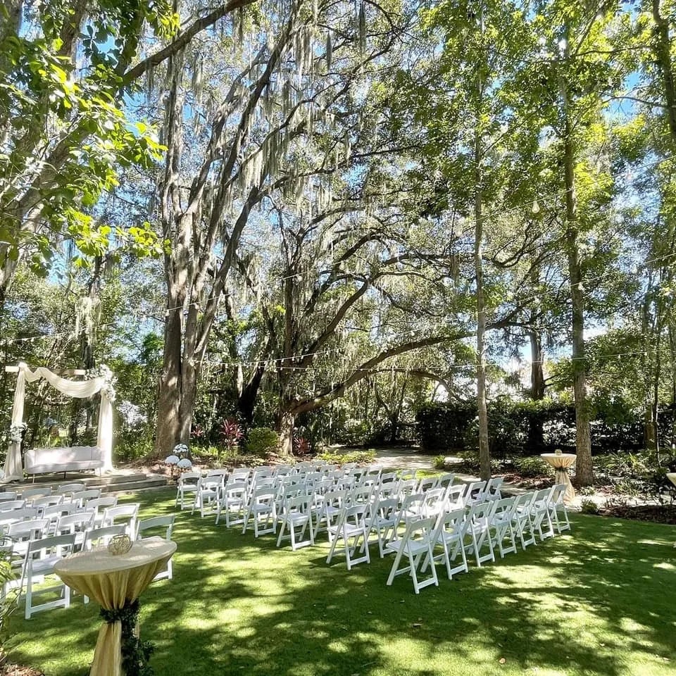 Outdoor wedding setup with white chairs and arch under trees with string lights
