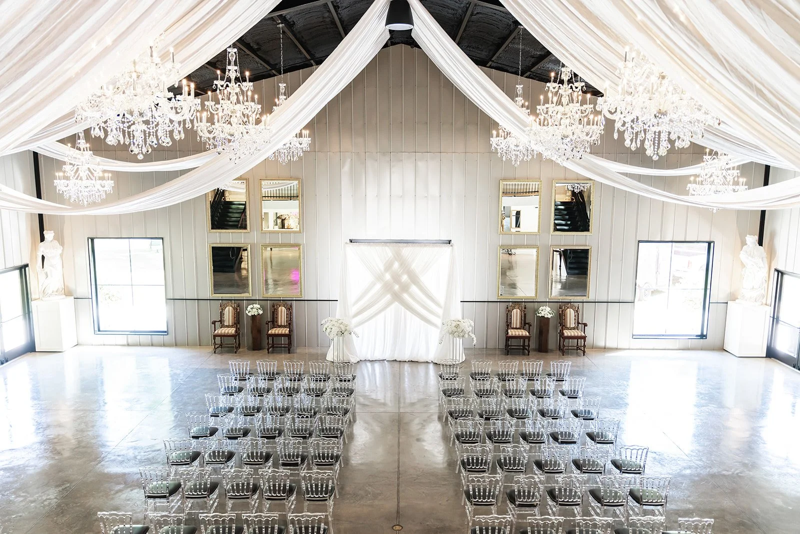 Elegant indoor wedding ceremony setup with sheer draped ceiling, crystal chandeliers, rows of clear chairs, and floral arrangements at the altar.