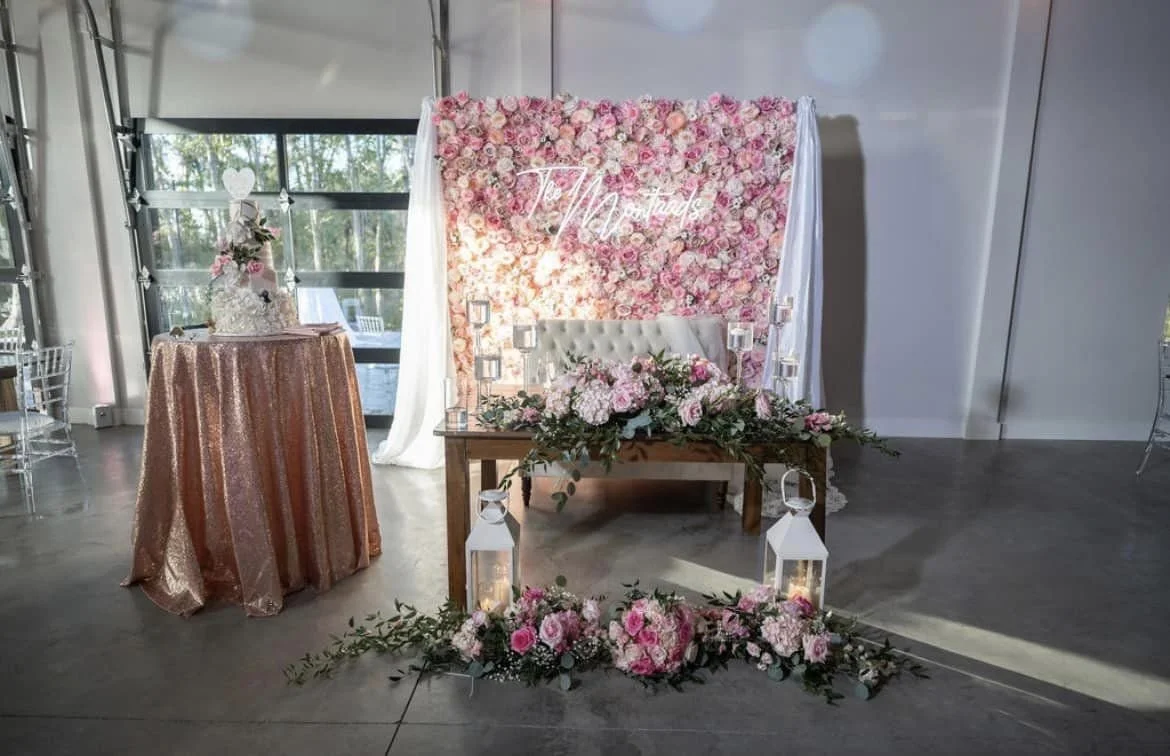 Wedding reception setup with floral backdrop, decorated table with lanterns, and a cake on a round table with a sequin tablecloth.