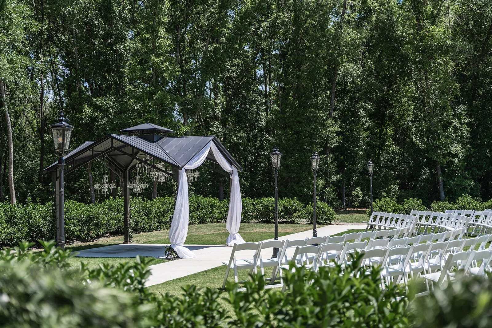 Outdoor wedding ceremony setup with white chairs and a decorated gazebo surrounded by greenery.