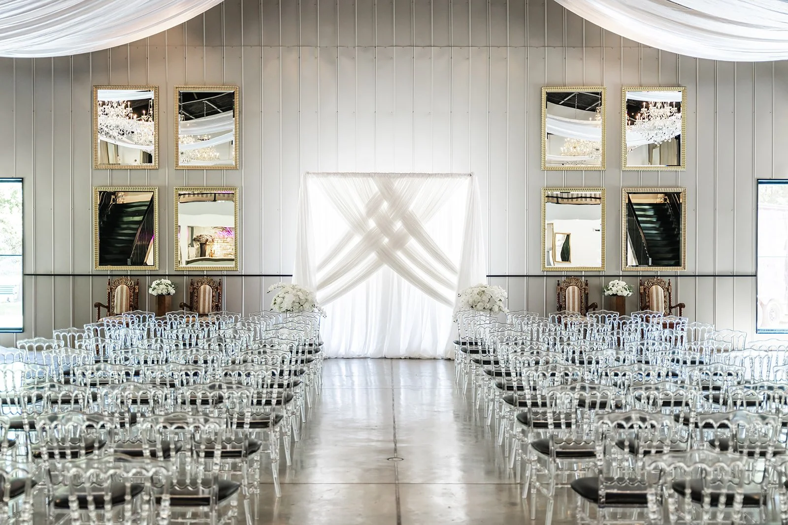 Elegant indoor wedding ceremony setup with transparent chairs, white aisle drapery, and mirrored wall decor.