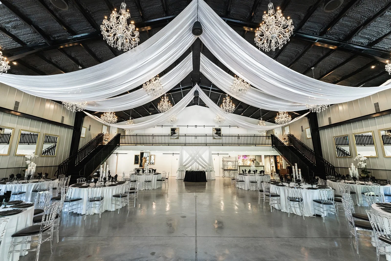 Elegant event hall with chandeliers, draped white fabric, round tables with white tablecloths, transparent chairs, and decorative centerpieces.