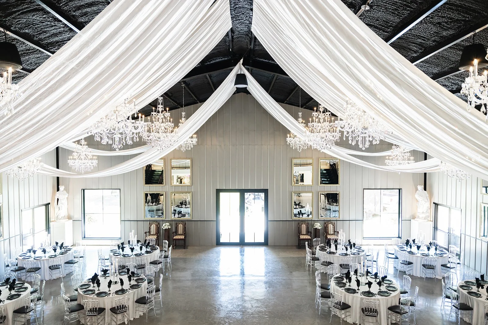 Elegant wedding reception venue with draped white fabric ceiling, chandeliers, round tables with white tablecloths and black accents, clear chairs, and large windows.