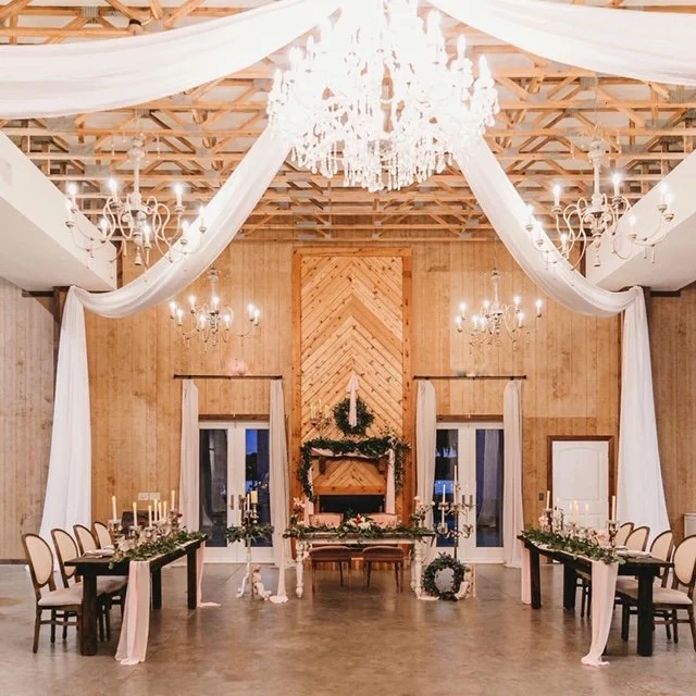 Elegant indoor wedding venue with chandeliers, draped white fabric, wooden wall panels, and decorated tables with candles and greenery.