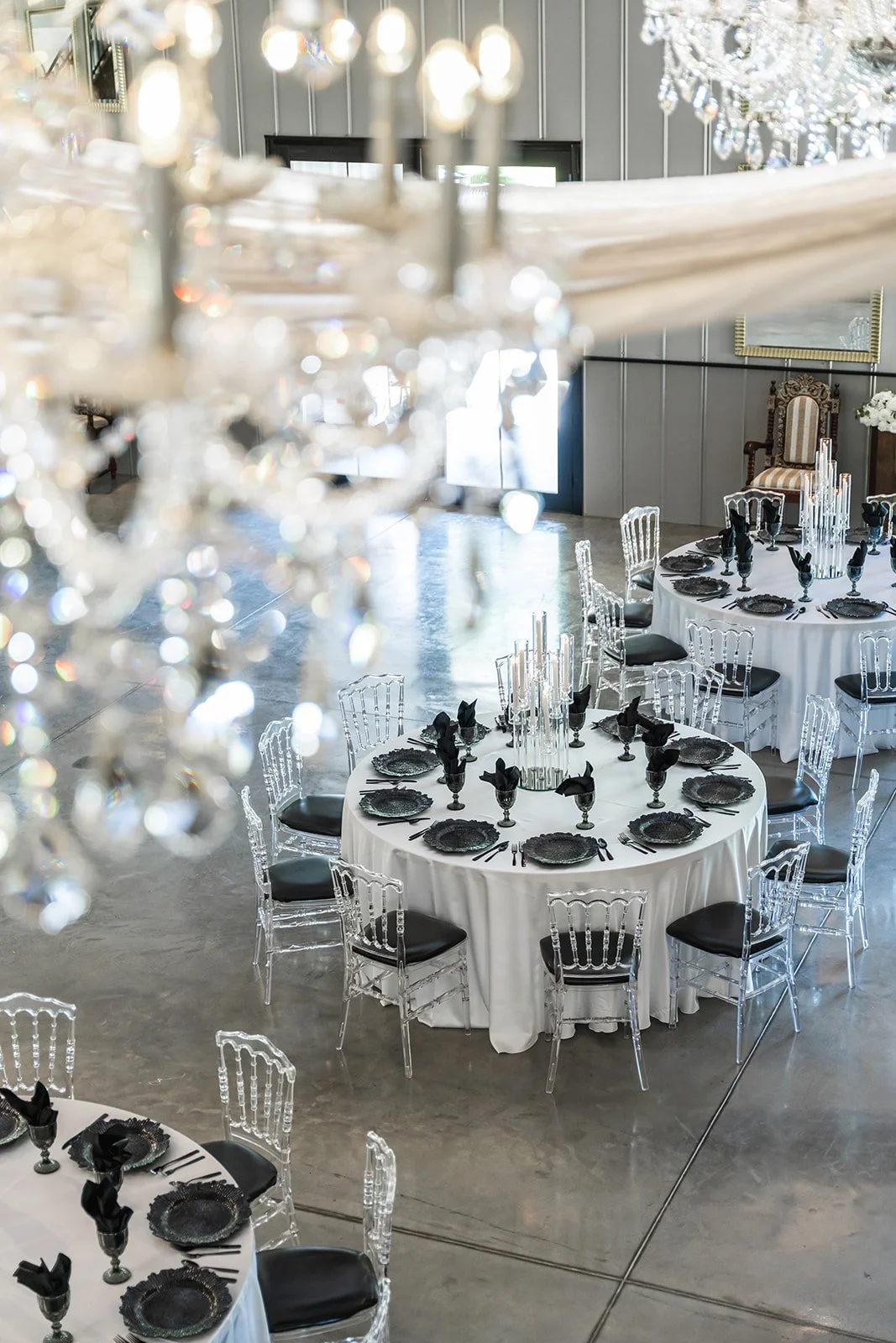 Elegant dining setup with round tables, white tablecloths, black plates, and clear chairs, viewed from above with chandelier crystals in the foreground.
