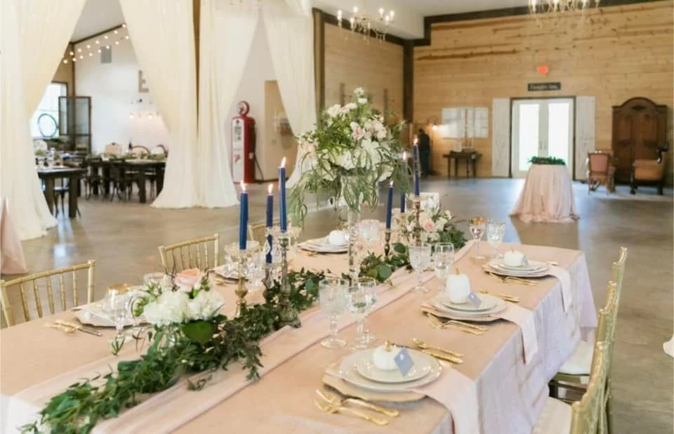 A elegantly decorated banquet hall with a long dining table set for an event. The table is draped with a pink tablecloth, adorned with floral centerpieces, greenery, and tall blue candles. Gold chiavari chairs are arranged around the table. The room 