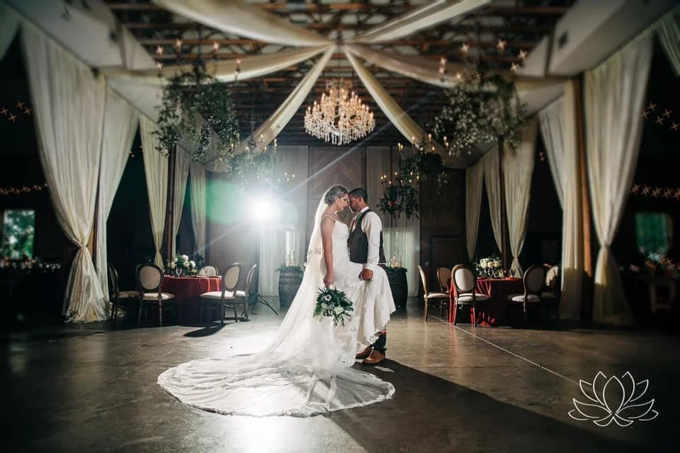 Bride and groom standing together in an elegantly decorated venue with draped curtains, chandeliers, and floral arrangements.