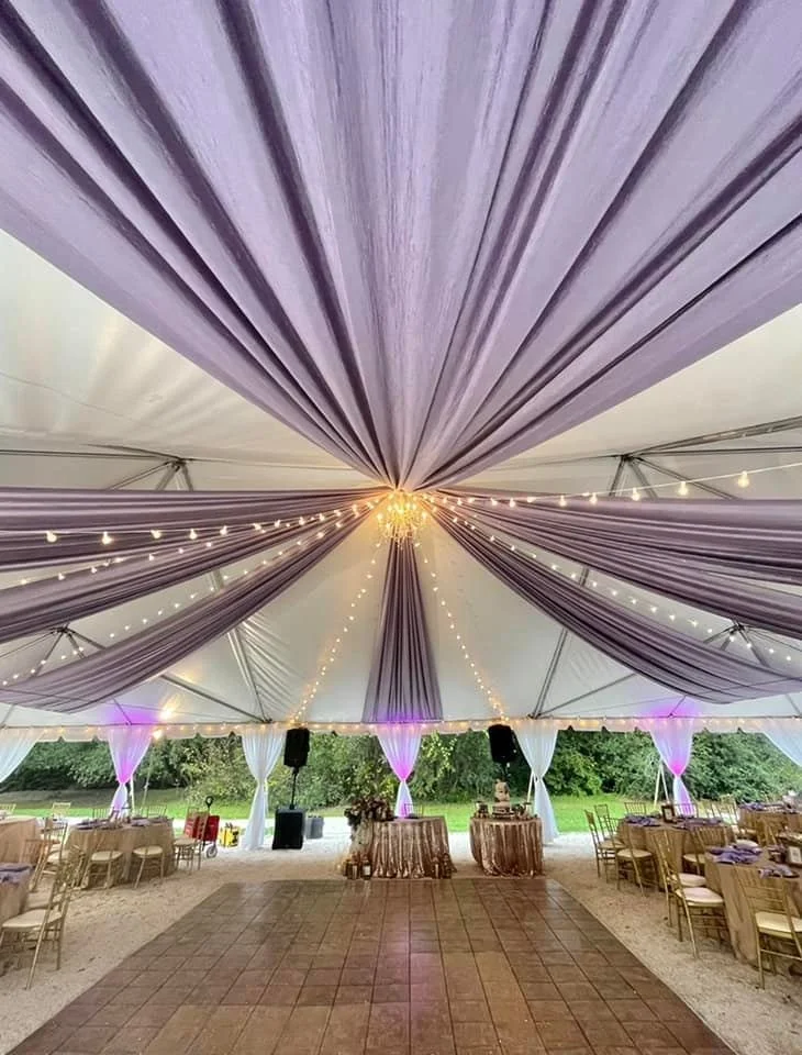 Interior of a tent with purple drapery and string lights, set up for an event with tables and chairs.