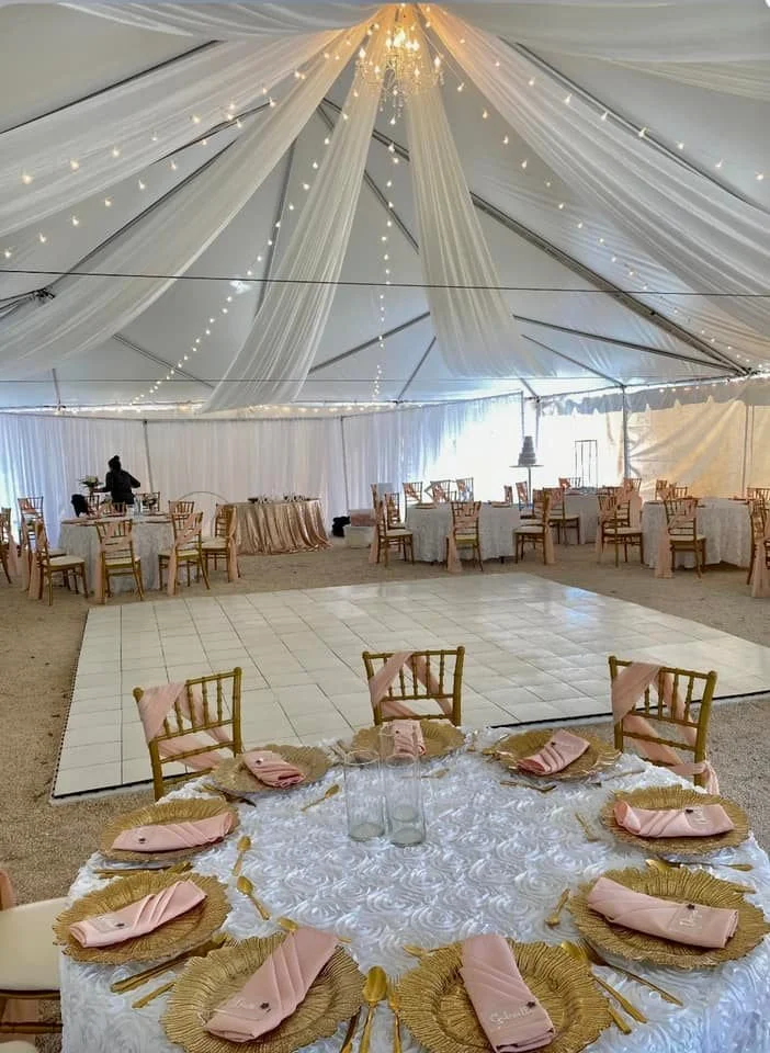 A wedding reception tent with elegant drapery, string lights, and a chandelier. The setup includes round tables with white tablecloths, gold chargers, pink napkins, and wooden chairs. A dance floor is centrally placed, surrounded by more tables and c