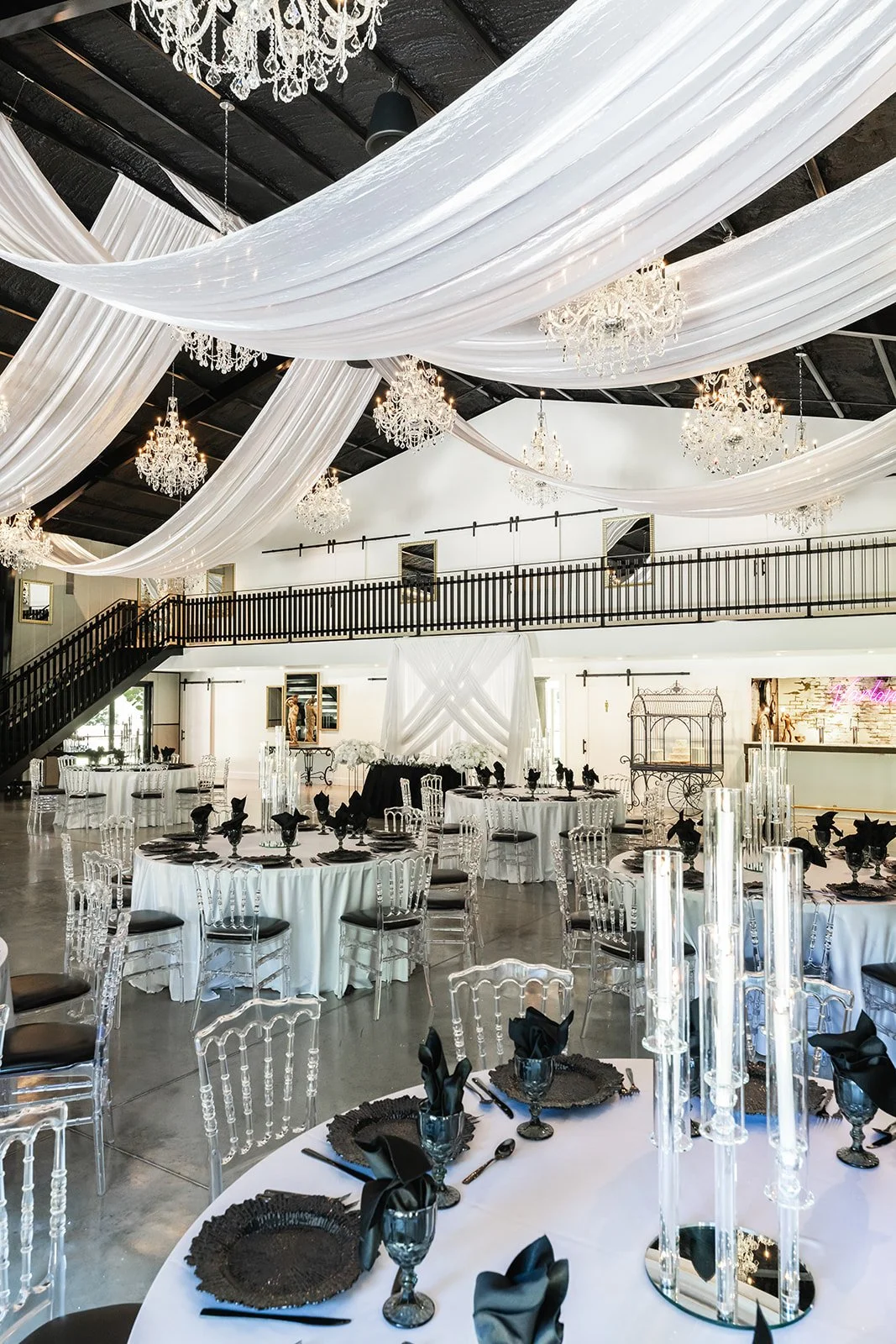 Elegant event venue interior with draped ceilings and chandeliers, round tables with white tablecloths, clear chairs, and black table settings.