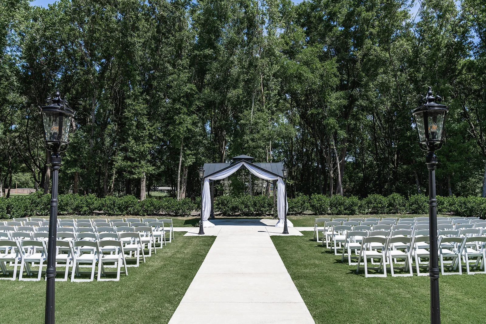 Outdoor wedding ceremony setup with white chairs arranged in rows, a draped archway, and two decorative lampposts, set on a grassy area with a backdrop of trees.