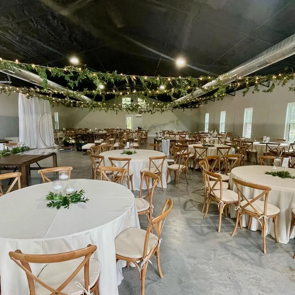Indoor event venue with round tables, white tablecloths, wooden chairs, greenery decor, and string lights on the ceiling.