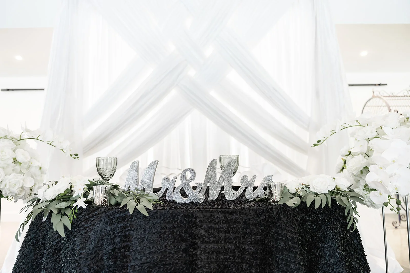 Wedding sweetheart table with "Mr & Mrs" sign, white floral arrangement, black tablecloth, and decorative white drapery backdrop.