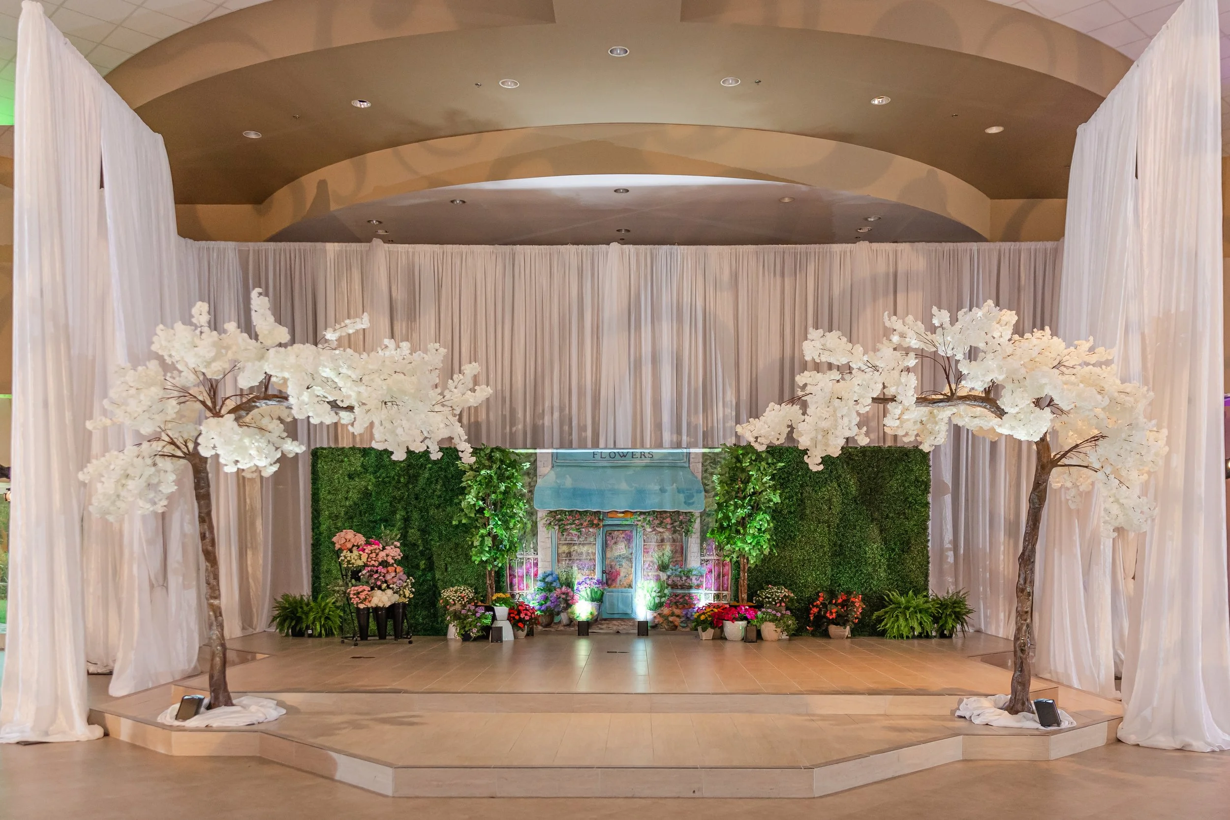 Decorated event stage with white artificial trees, draped curtains, floral arrangements, and a backdrop resembling a small flower shop entrance.