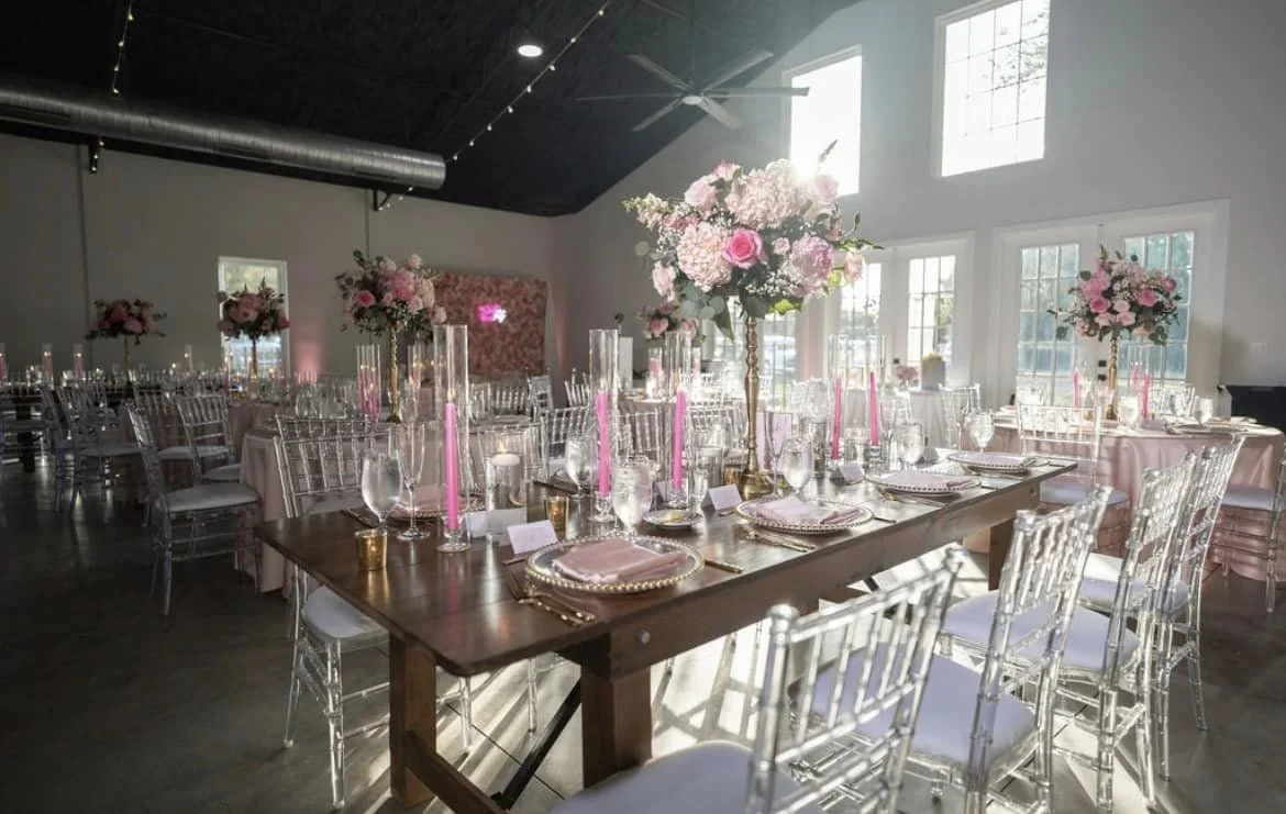 Elegant wedding reception setup with floral centerpieces, pink candles on long tables, clear chairs, and natural lighting in a spacious venue.