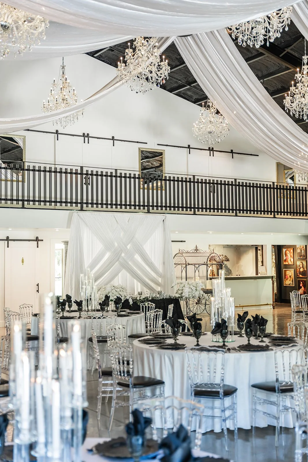 Elegant banquet hall with draped ceiling, crystal chandeliers, round tables set with black and white decor, and transparent chairs.