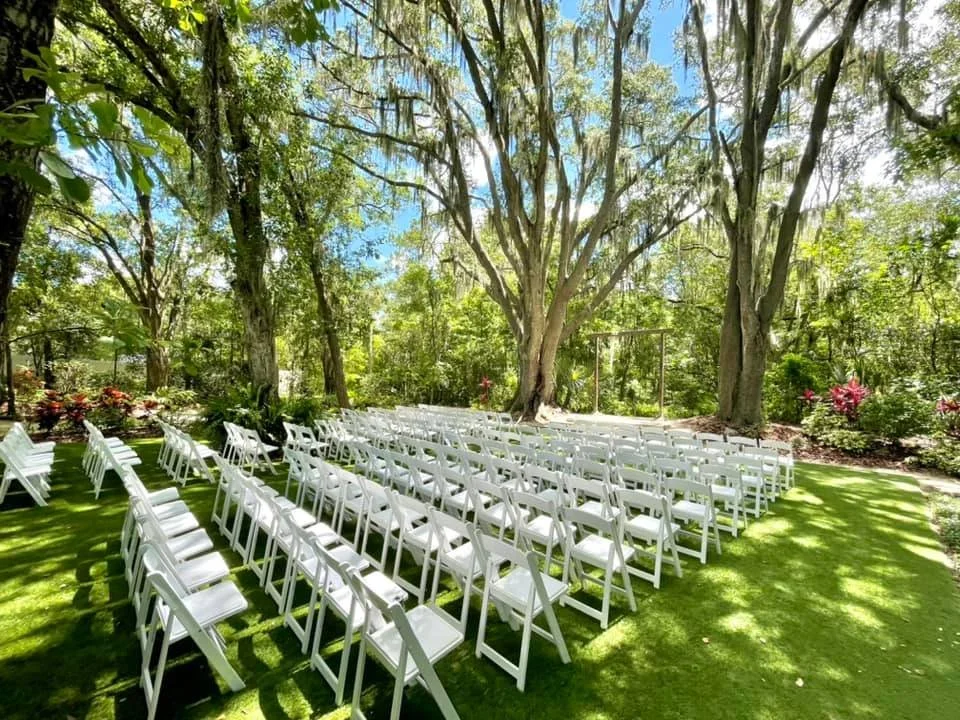Outdoor wedding ceremony setup with white folding chairs arranged on a grassy area surrounded by trees and greenery.