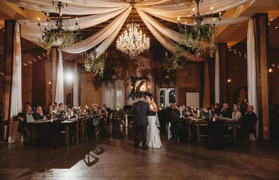Couple dancing at a wedding reception in an elegantly decorated venue with chandeliers and draped fabric. Guests seated at tables.