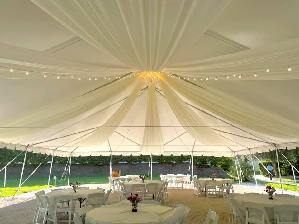 Large outdoor tent with white draping and string lights, round tables with white chairs and floral centerpieces.