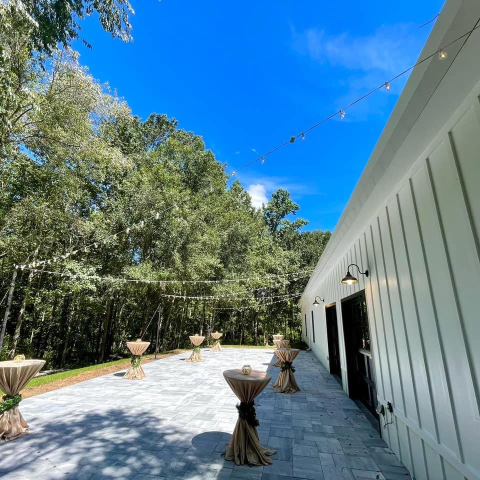 Outdoor event space with cocktail tables, string lights, and trees, adjacent to a white building under a clear blue sky.