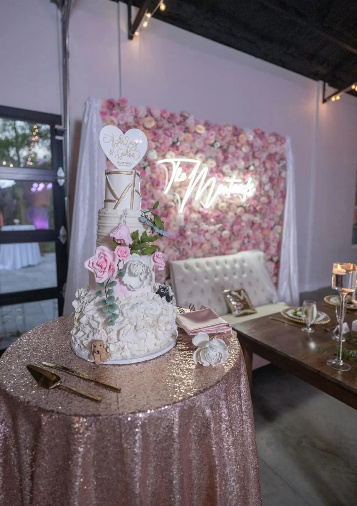 Wedding cake on a sequin tablecloth with floral decorations and a neon sign backdrop.