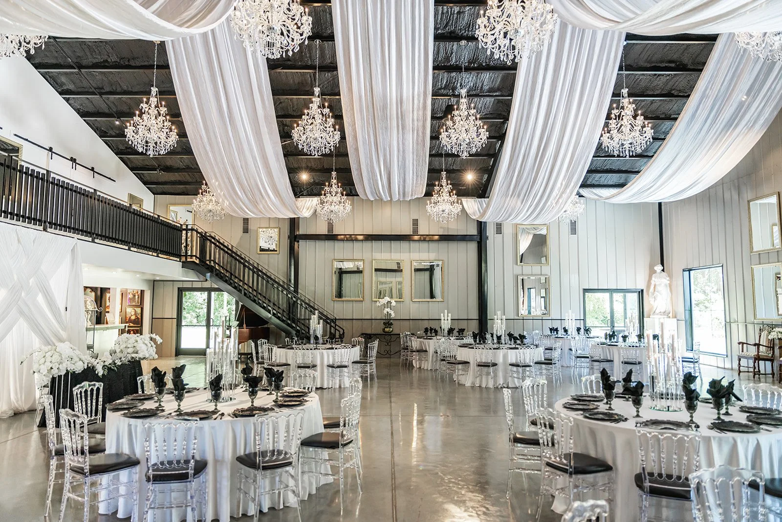 Elegant wedding reception hall with white drapes, chandeliers, round tables with white tablecloths, black napkins, and transparent chairs.