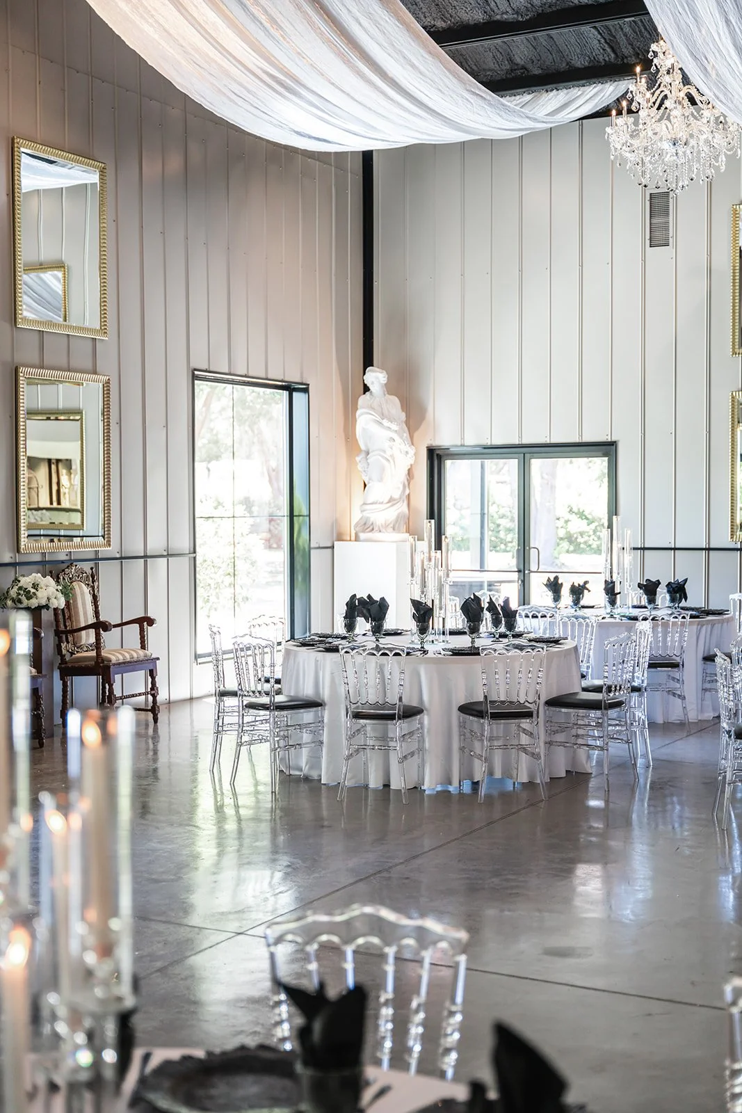 Elegant banquet hall with round tables, black napkins, transparent chairs, chandeliers, mirrors, and a white statue.