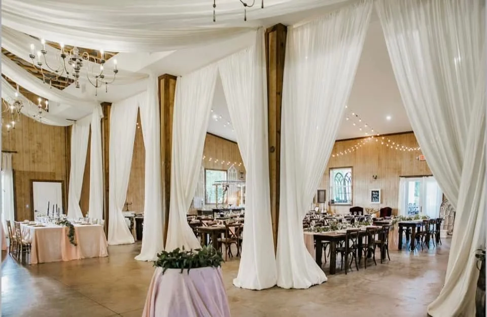 Elegant indoor event venue with white draped curtains and wooden beams, featuring tables set for a formal gathering.