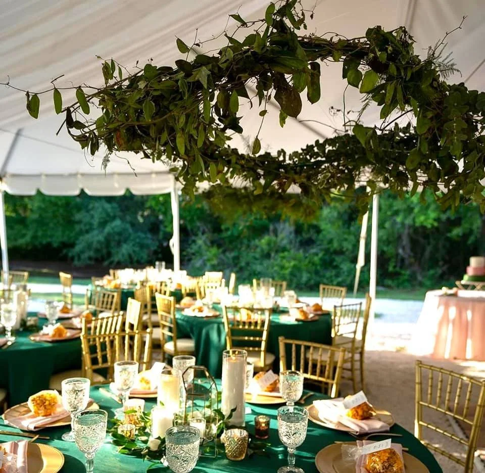 Outdoor wedding reception setup with round tables covered in green tablecloths, gold chairs, crystal glassware, and decorative centerpieces under a tent. A hanging greenery wreath is overhead, and a wedding cake is visible in the background.