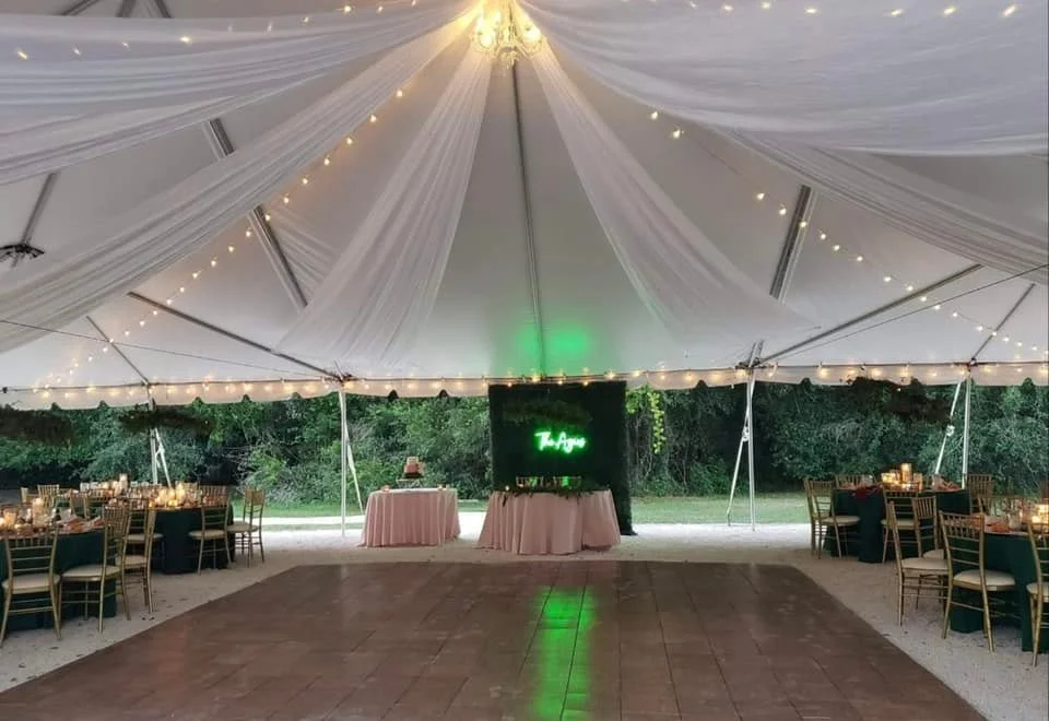 Outdoor wedding reception tent with draped fabric and string lights, tables with candles and chairs, and a neon sign on a greenery backdrop.