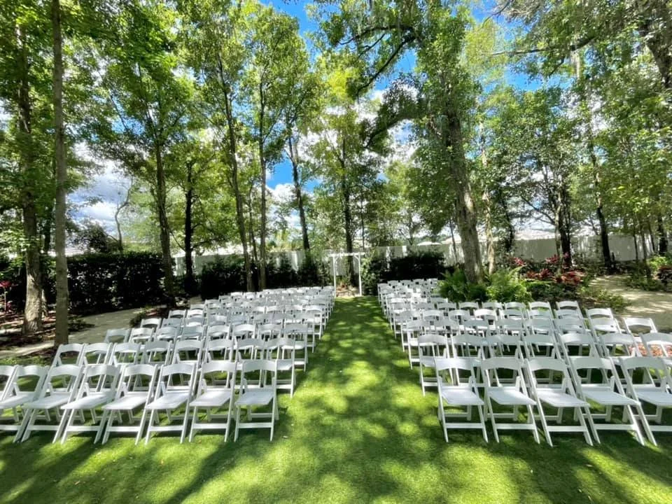 Outdoor wedding ceremony setup with rows of white chairs on grass, surrounded by trees, and a white arbor at the end.