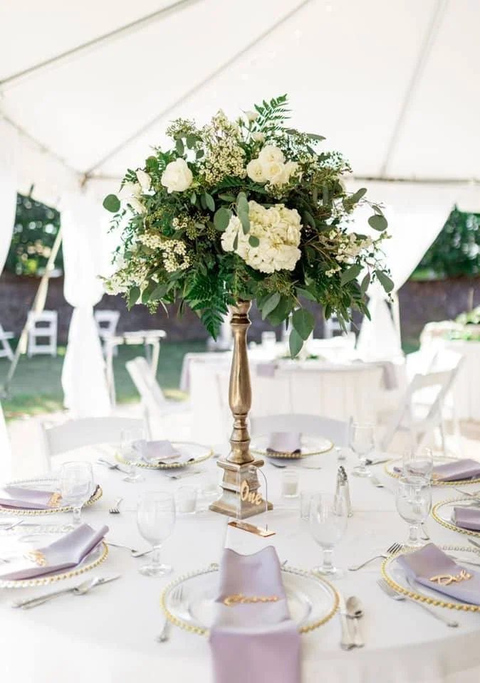 Elegant wedding reception table setting with a tall floral centerpiece, white tablecloth, lavender napkins, and gold accents under a white tent.